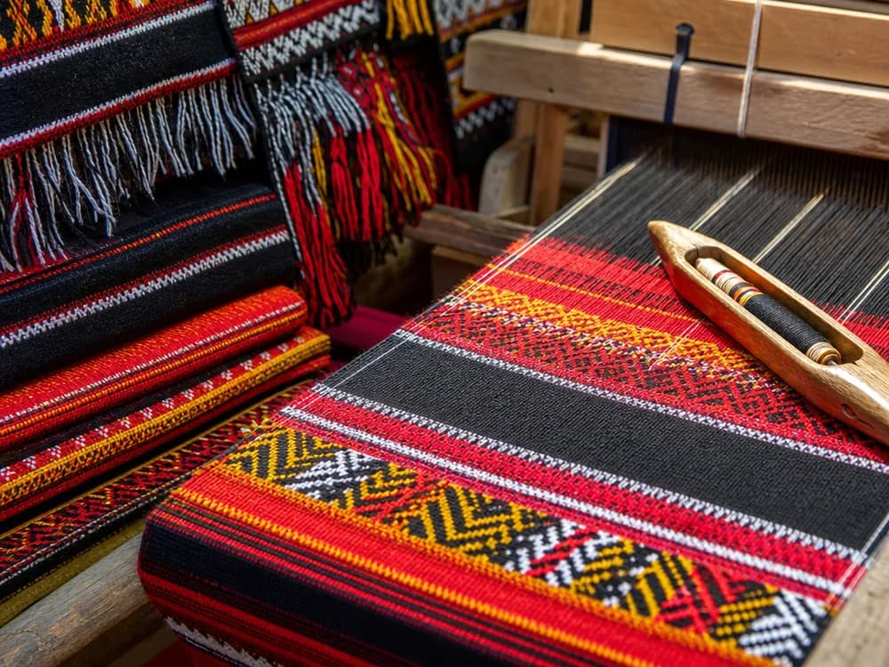 Brightly coloured handwoven shawls with red and black geometric patterns stacked in sunlight