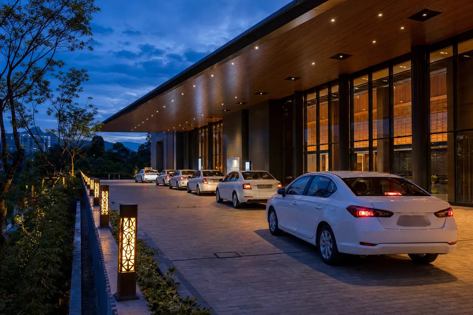 A line of white sedans on the lit drive of a convention centre at dusk